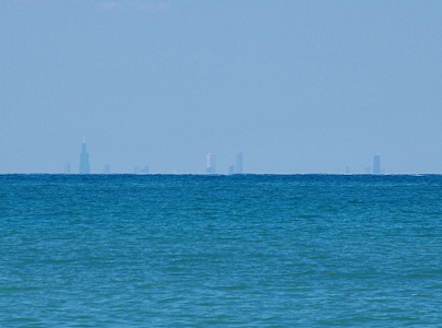 [A lake view with four very tall buildings and a number of smaller ones visible behind the haze at the water's surface.]